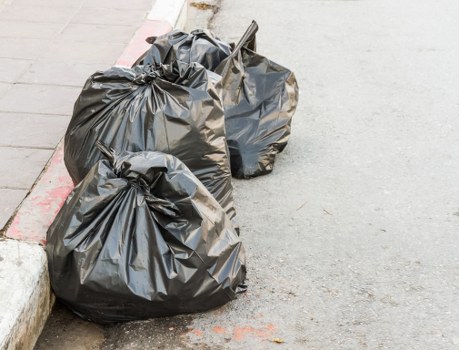 Three black refuse bags, made from thick, glossy plastic, are placed on a concrete pavement near the kerbside, partially resting against a curbstone; they appear to be filled with household or garden waste, with some slightly bulging and tied at the top. The bags are situated on a grey, slightly rough-textured street surface, with a faint red line marking the edge of a parking bay or roadside boundary visible behind them. The background features a small section of a light-coloured pavement edge, possibly part of a residential or commercial area in Hayes, suggesting a typical rubbish collection scene. The presence of the waste bags indicates a rubbish removal or clearance activity carried out by a local service such as Garden Clearance Hayes, catering to the area's waste disposal needs.