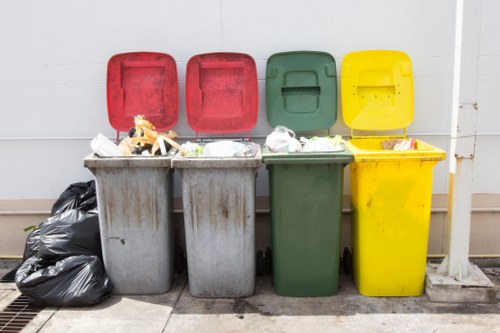 The image displays four outdoor rubbish bins positioned in a row against a white wall, each with a distinct coloured lid: red, red, green, and yellow, from left to right. The grey bins beneath the red lids contain various waste items, including plastic packaging and paper, with some overflow visible on the tops. To the far left, a black trash bag is placed on the ground, resting against the grey bin. The green and yellow bins are closed, with no visible contents. The ground surface appears to be paved concrete, and a metal drain grate is visible at the bottom left corner of the image. The scene suggests an outdoor urban or residential setting, typically associated with waste collection and rubbish removal services, such as those provided by Garden Clearance Hayes in Hayes, Middlesex, supporting local rubbish disposal needs.