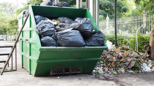 A large green skip bin filled with black refuse bags is positioned on an outdoor pavement, adjacent to a garden area edged by a metal fence and trees. The bags appear to contain garden waste or debris, with some close to overflowing and stacked neatly inside the bin. To the right, there is a small pile of mixed garden rubbish, including leaves, cardboard, and other organic or household waste, scattered on the ground. The background features a pathway with some greenery, trees, and nearby residential hedges or bushes, indicative of a suburban setting. The scene is lit by natural daylight, emphasizing the textures of the bags, the bin's smooth surface, and the surrounding environment, consistent with a typical waste collection or rubbish removal service, such as those offered by Garden Clearance Hayes in Hayes or nearby areas.