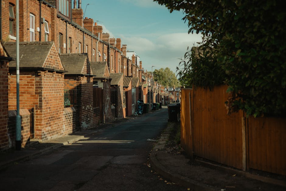A narrow residential street lined with red-brick terraced houses featuring pitched roofs and chimney stacks. The houses have small front yards with brick walls and a mix of windows, some with green or